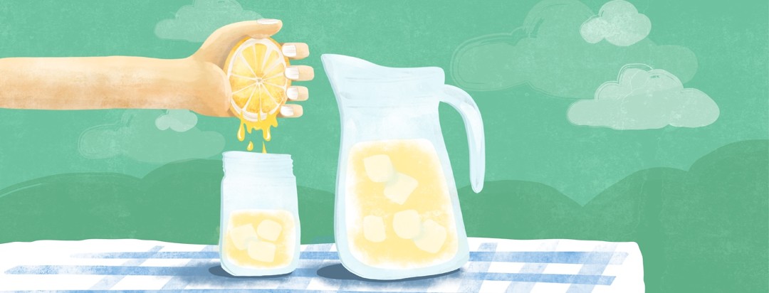A hand squeezing a lemon into a glass next to a pitcher of lemonade.