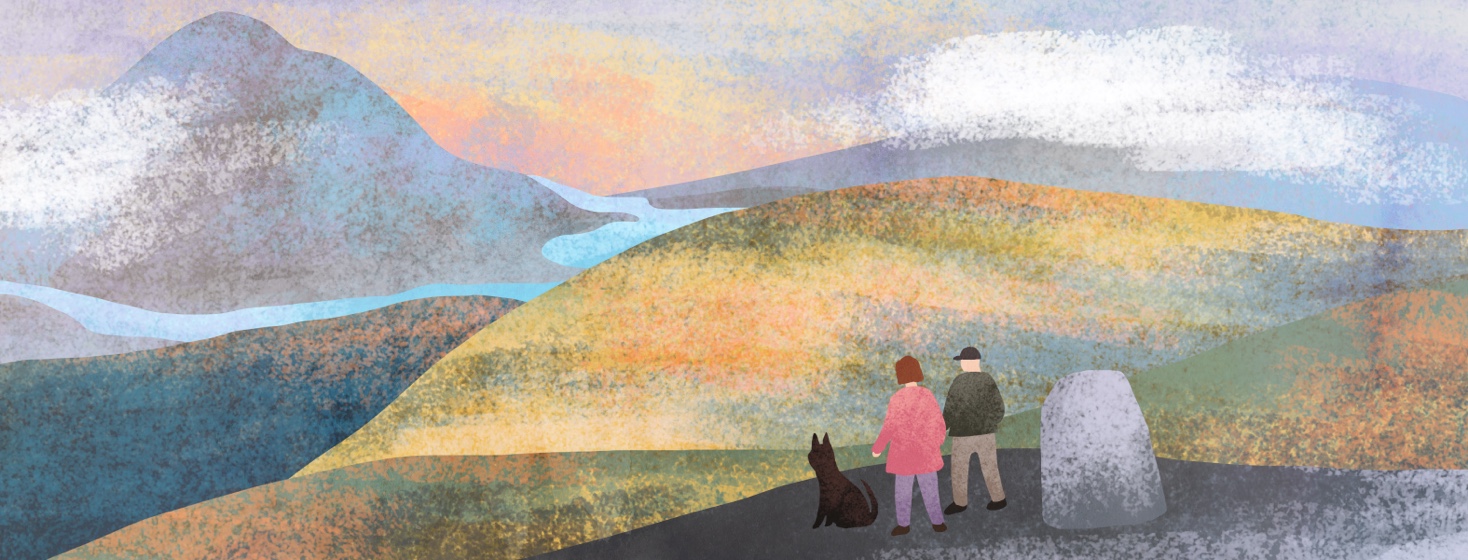 A man, woman and dog on a hike look out over a hill towards a vast green landscape with mountains and rivers.