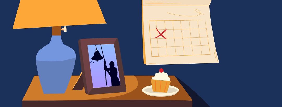 A table with a lamp has a picture framed of someone ringing a bell while a cupcake and calendar sit nearby.